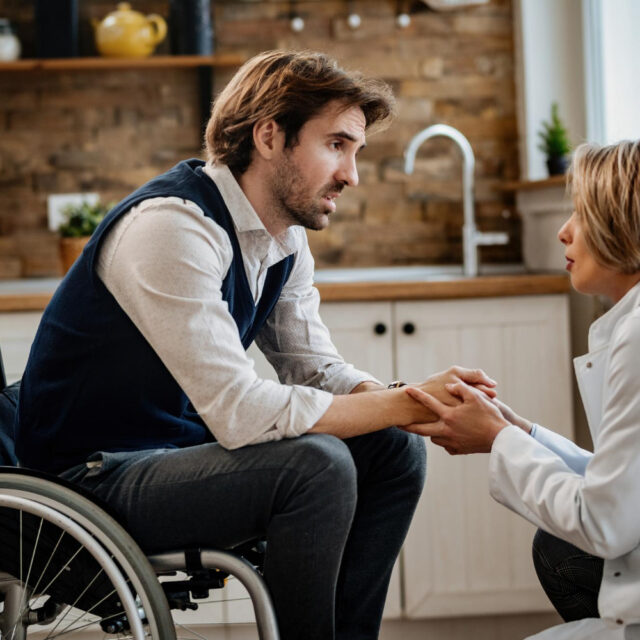 Young Distraught Man Wheelchair Holding Hands With Doctor While Talking Her During Home Visit